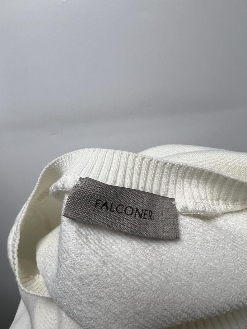 May include: Close-up of a white sweater with a ribbed neckline. A gray label with the brand name "FALCONERI" is sewn on the sweater. The sweater is made of a soft, textured material.