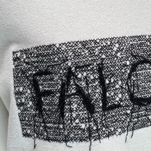 May include: White sweatshirt with a black, open-weave patch. The patch features the word "FALL" in a stylized font. The sweatshirt has a simple, minimalist design, with the text as the focal point. The fabric appears to be a soft, comfortable material.