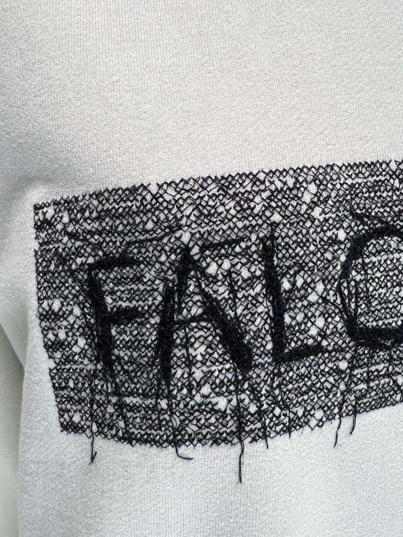 May include: White sweatshirt with a black, open-weave patch. The patch features the word "FALL" in a stylized font. The sweatshirt has a simple, minimalist design, with the text as the focal point. The fabric appears to be a soft, comfortable material.