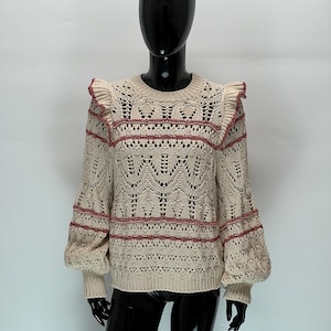 May include: Cream-colored knit sweater with a round neck and long sleeves. The sweater features a detailed openwork design with red accents on the shoulders and sleeves. The sleeves have a slight puff at the elbow and ribbed cuffs.