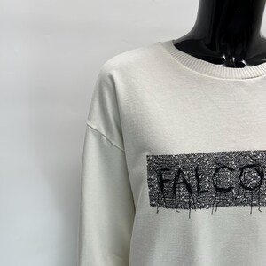 May include: Off-white sweatshirt with the word "FALCO" in a black, textured rectangle. The sweatshirt has a crew neck and long sleeves. The text appears to be embroidered or printed on the front.