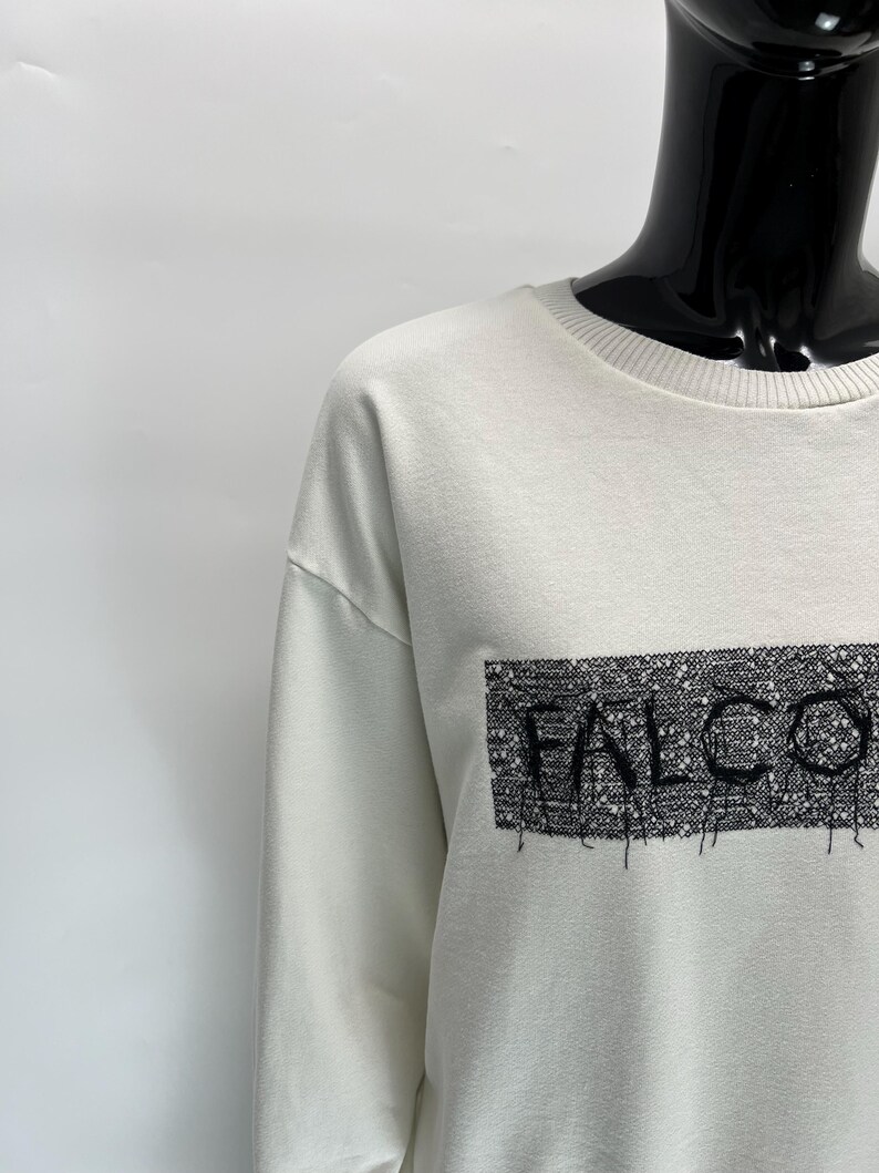 May include: Off-white sweatshirt with the word "FALCO" in a black, textured rectangle. The sweatshirt has a crew neck and long sleeves. The text appears to be embroidered or printed on the front.
