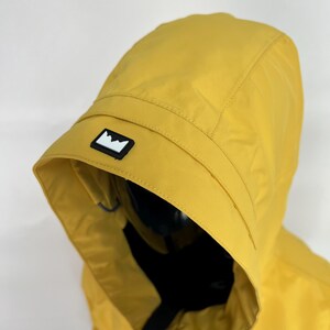 May include: Mustard yellow hooded jacket with a black and white mountain logo on the hood. The jacket has a black zipper and a black interior. The hood is up, and the jacket appears to be made of a lightweight, water-resistant material.