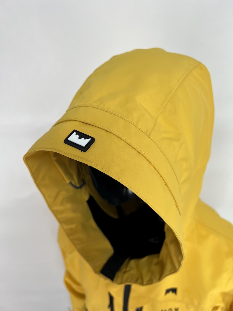 May include: Mustard yellow hooded jacket with a black and white mountain logo on the hood. The jacket has a black zipper and a black interior. The hood is up, and the jacket appears to be made of a lightweight, water-resistant material.