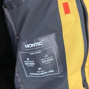 May include: Close-up of a black and yellow jacket interior. A tag reads "MONTEC MOUNTAIN TECHNOLOGY SHELLFABRIC" with details on waterproof and breathability ratings. The jacket features a zipper and a small red accent.