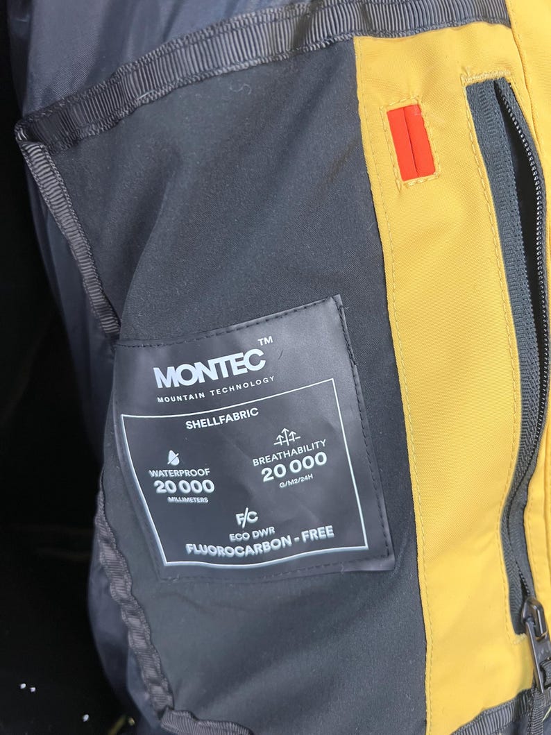 May include: Close-up of a black and yellow jacket interior. A tag reads "MONTEC MOUNTAIN TECHNOLOGY SHELLFABRIC" with details on waterproof and breathability ratings. The jacket features a zipper and a small red accent.