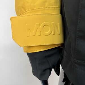 May include: Close-up of a yellow jacket sleeve with the embossed letters "MON" on the cuff. The jacket is paired with black gloves and a black garment. The jacket's bright color contrasts with the dark gloves.