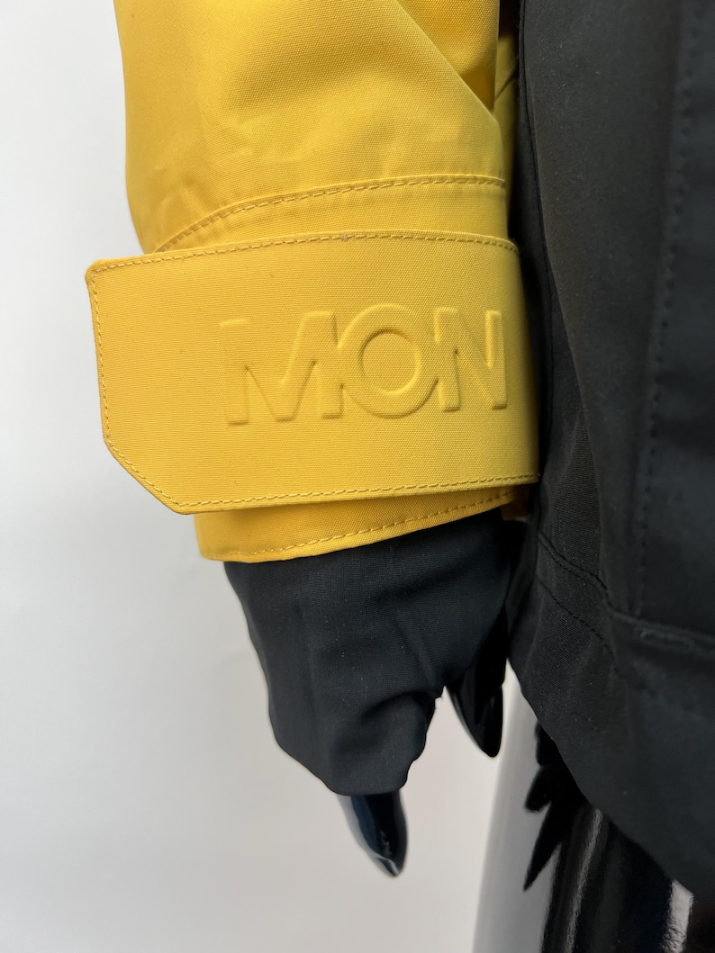 May include: Close-up of a yellow jacket sleeve with the embossed letters "MON" on the cuff. The jacket is paired with black gloves and a black garment. The jacket's bright color contrasts with the dark gloves.