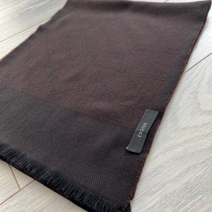 May include: A brown scarf with a darker brown border and fringe. A black tag with the word "GUCCI" is attached to the scarf. The scarf is made of a soft material.