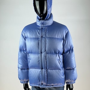 Puede incluir: Una chaqueta acolchada azul claro con capucha. La chaqueta tiene un acabado brillante y está acolchada con líneas horizontales.