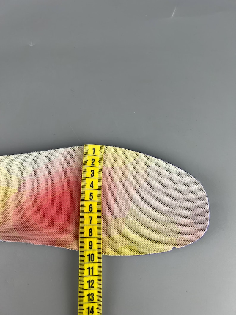 May include: A colorful shoe insole with a gradient of pink, yellow, and white. A yellow measuring tape is placed next to the insole, showing a measurement of approximately 5 inches.