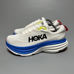 Puede incluir: Una zapatilla deportiva Hoka blanca con una entresuela gruesa y superpuesta en blanco y azul, marca Hoka negra en el lateral, detalles en amarillo neón en los ojales y una pequeña lengüeta roja en el talón. Parte superior de malla.