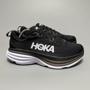 Könnte beinhalten: Paar schwarze Laufschuhe mit weißen und braunen Sohlen. Das Wort "HOKA" ist weiß auf der Seite aufgedruckt. Die Schuhe haben schwarze Schnürsenkel und eine schwarze Zuglasche an der Ferse.