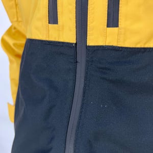 May include: A yellow and black jacket with a full-length zipper. The jacket has a color-block design, with yellow on the upper portion and black on the lower. The jacket has three vertical black stripes on the yellow section.