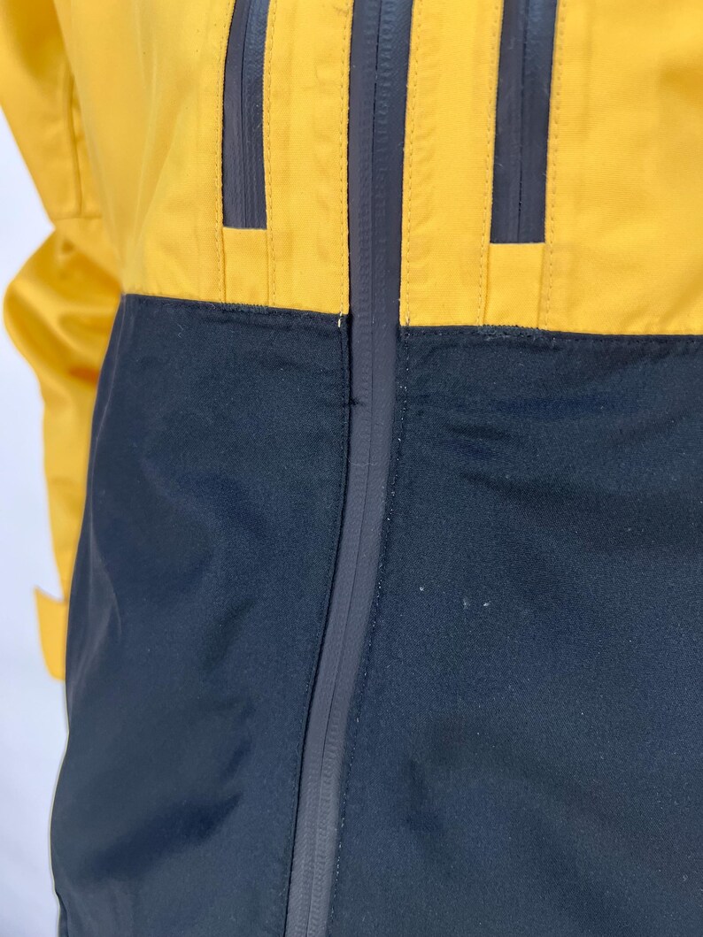 May include: A yellow and black jacket with a full-length zipper. The jacket has a color-block design, with yellow on the upper portion and black on the lower. The jacket has three vertical black stripes on the yellow section.