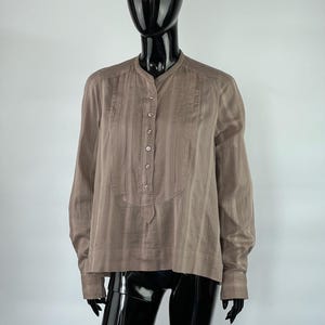 May include: A long-sleeved, taupe-colored shirt with a mandarin collar and a button-down placket. The shirt has vertical striped detailing and is displayed on a black mannequin. The sleeves have buttoned cuffs.