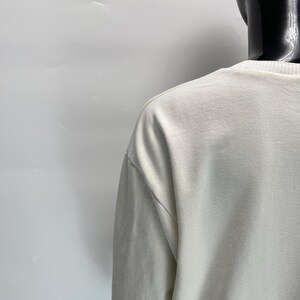 May include: Off-white long-sleeve shirt with a crew neck. The shirt is displayed on a black mannequin against a white background. The fabric appears to be a soft, textured material.