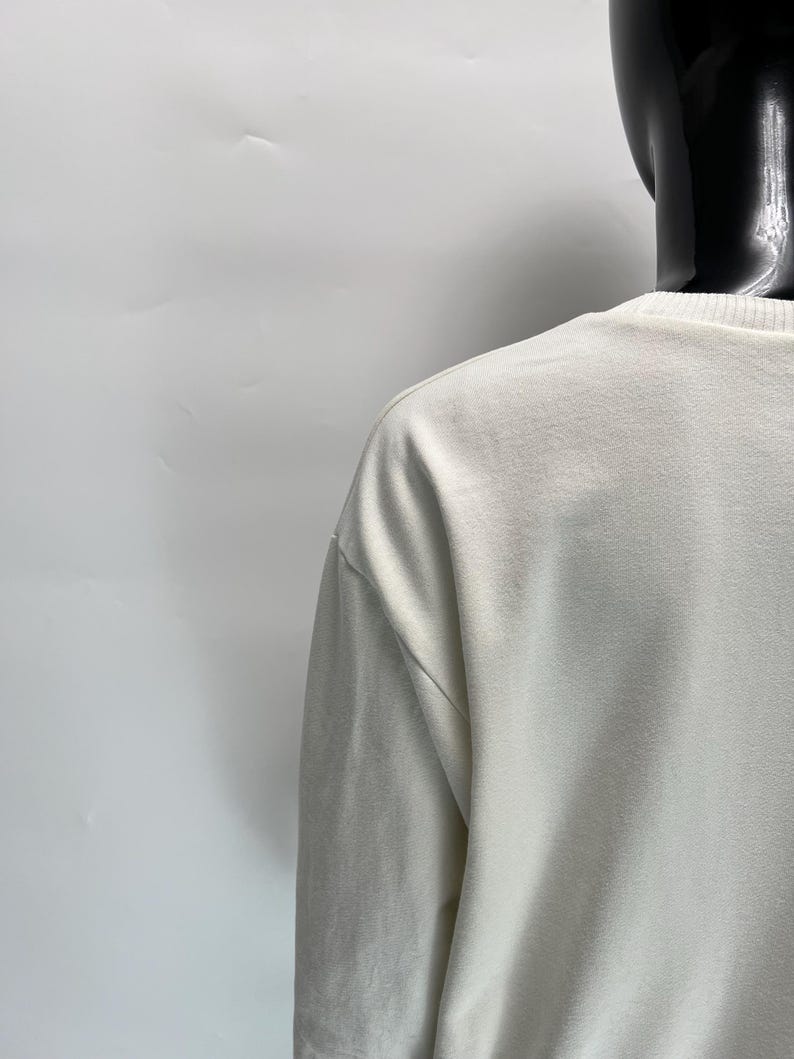 May include: Off-white long-sleeve shirt with a crew neck. The shirt is displayed on a black mannequin against a white background. The fabric appears to be a soft, textured material.
