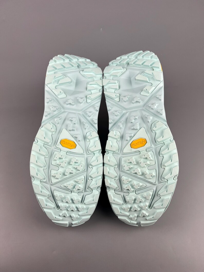 May include: A pair of light blue hiking shoes, soles facing up, with a textured pattern and the brand name "Vibram" in yellow. The shoes are designed for outdoor activities and provide grip and support.