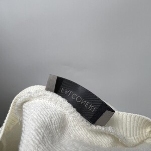 May include: Close-up of a cream-colored, textured fabric with a black label attached. The label has white text that reads "EVICOMEBI". The fabric appears to be a sweater or sweatshirt material.