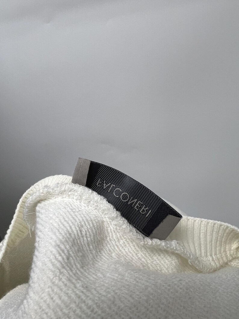 May include: Close-up of a cream-colored, textured fabric with a black label attached. The label has white text that reads "EVICOMEBI". The fabric appears to be a sweater or sweatshirt material.