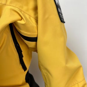 May include: Close-up of a bright yellow jacket with a black zipper and a black and white patch on the sleeve. The patch features a mountain graphic and the text "MONT TEC". The jacket has a black panel on the side.