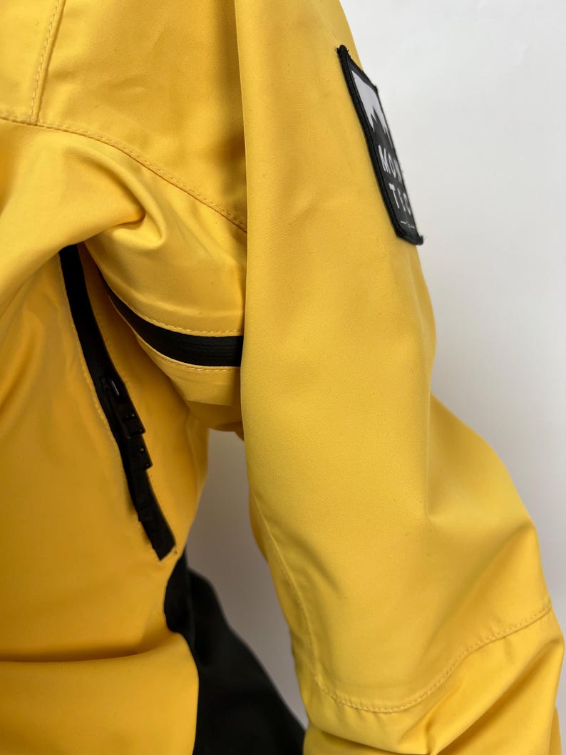 May include: Close-up of a bright yellow jacket with a black zipper and a black and white patch on the sleeve. The patch features a mountain graphic and the text "MONT TEC". The jacket has a black panel on the side.