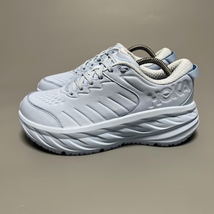 Op de afbeelding: Paar lichtblauwe Hoka hardloopschoenen. De schoenen hebben een dikke, witte, gebogen zool en een lichtblauw bovenwerk met het Hoka-logo in reliëf aan de zijkant. De veters en binnenvoering zijn lichtgrijs.