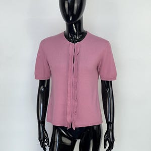May include: A short-sleeved, dusty rose cardigan with a ruffled placket down the front. The top is displayed on a black mannequin against a white background. The cardigan has a round neckline.