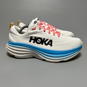 Puede incluir: Zapatillas deportivas blancas con texto "HOKA" en negro y cordones color coral. La suela presenta un diseño en capas con detalles en blanco, azul claro y amarillo. Las zapatillas están diseñadas para correr y otras actividades deportivas.