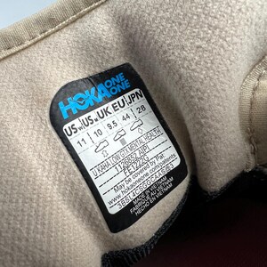 May include: Close-up of the inside of a beige Hoka One shoe. A black and blue label displays the brand name and shoe size, US 11, UK 10, EU 44, and JPN 28. The label also includes the style name, U KAHA LOW GTX MENTAL HEALTH.