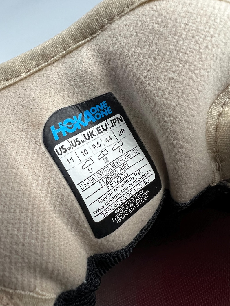 May include: Close-up of the inside of a beige Hoka One shoe. A black and blue label displays the brand name and shoe size, US 11, UK 10, EU 44, and JPN 28. The label also includes the style name, U KAHA LOW GTX MENTAL HEALTH.