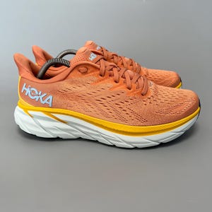 Puede incluir: Par de zapatillas de running naranjas con suelas blancas y amarillas. Las zapatillas tienen el nombre de la marca "HOKA" en letras azules en el lateral. Diseñadas para actividades deportivas, cuentan con una parte superior de malla transpirable.