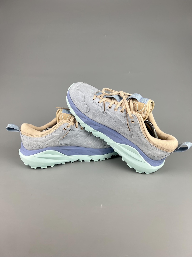 May include: A pair of athletic shoes in a light gray color with light blue and tan accents. The soles are light green. The shoes are designed for outdoor activities and have a textured upper.