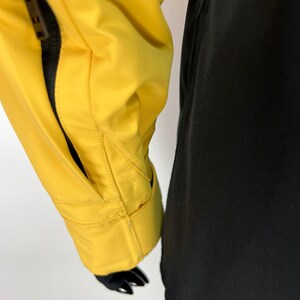 May include: Close-up of a yellow jacket sleeve with a black sleeve next to it. The yellow jacket has a zippered pocket and a black lining. The jacket is made of a smooth, possibly waterproof material.
