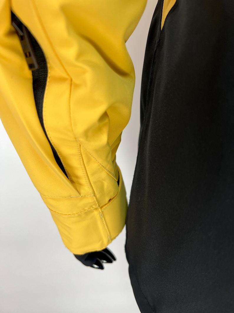 May include: Close-up of a yellow jacket sleeve with a black sleeve next to it. The yellow jacket has a zippered pocket and a black lining. The jacket is made of a smooth, possibly waterproof material.