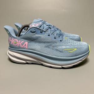 Puede incluir: Par de zapatillas para correr Hoka azul claro. Las zapatillas tienen una entresuela blanca, una parte superior azul claro con la palabra "HOKA" en rosa y un acento amarillo. Diseñadas para correr y actividades atléticas.