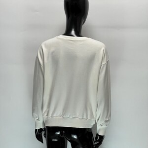 May include: A cream-colored sweatshirt with long sleeves and a ribbed hem and cuffs. The back view of the sweatshirt is displayed on a black mannequin against a white background. The sweatshirt has a crew neck.