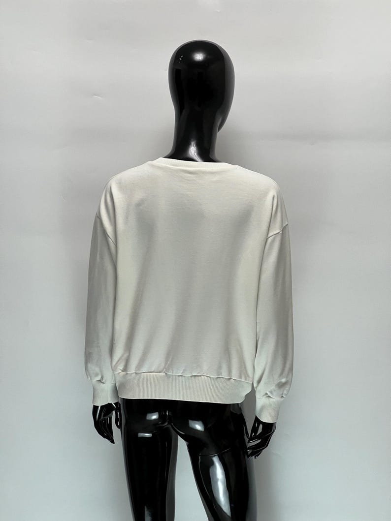 May include: A cream-colored sweatshirt with long sleeves and a ribbed hem and cuffs. The back view of the sweatshirt is displayed on a black mannequin against a white background. The sweatshirt has a crew neck.