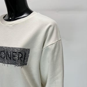 May include: Off-white sweatshirt with the word "ONERI" in black, stylized text. The text is within a rectangular design with a textured pattern. The sweatshirt has a crew neck and long sleeves.