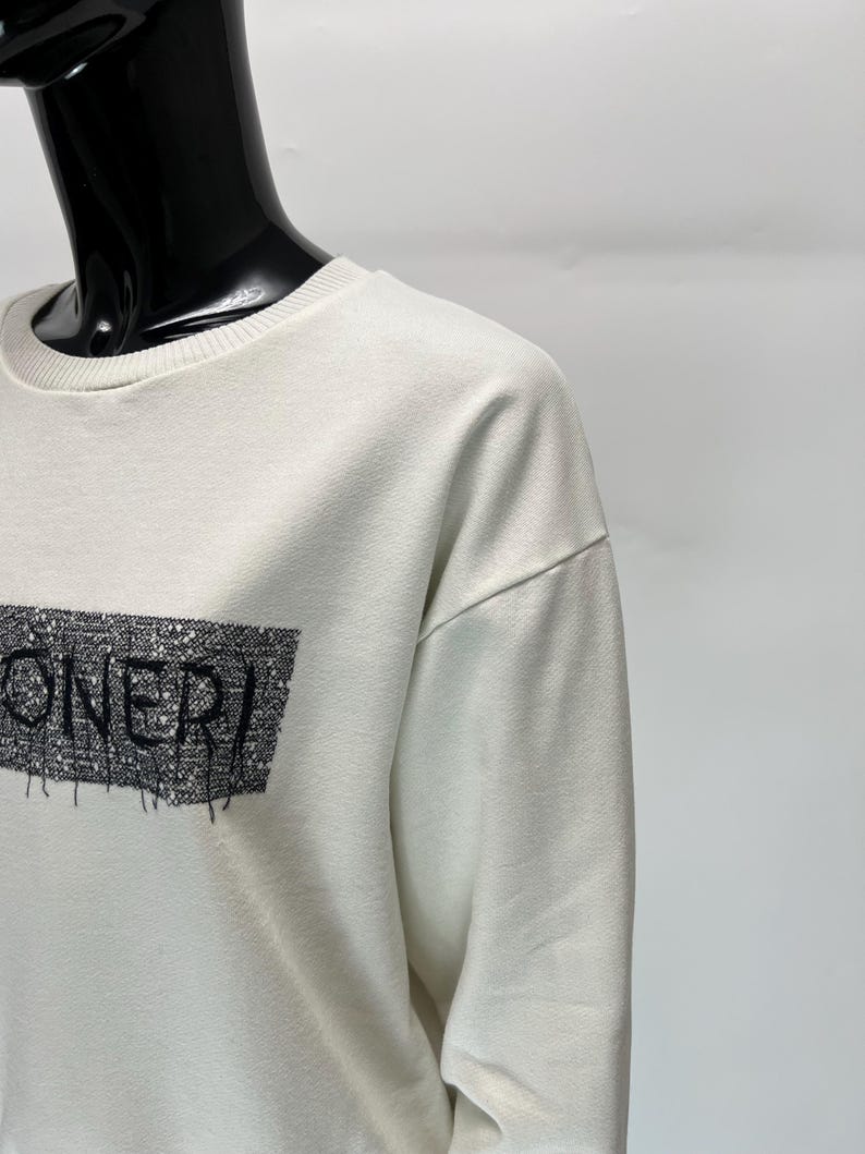 May include: Off-white sweatshirt with the word "ONERI" in black, stylized text. The text is within a rectangular design with a textured pattern. The sweatshirt has a crew neck and long sleeves.