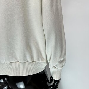 May include: A light cream-colored sweatshirt with ribbed cuffs and hem. The long sleeves have a relaxed fit, and the fabric appears soft. The garment is displayed on a black mannequin.