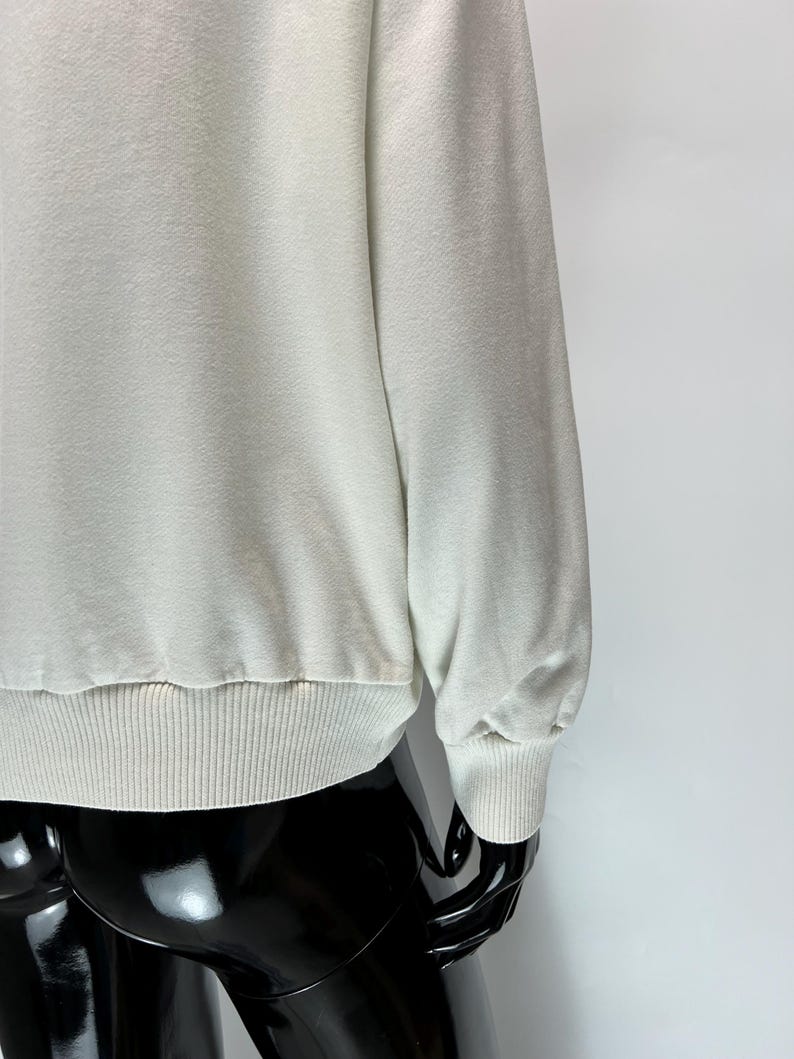 May include: A light cream-colored sweatshirt with ribbed cuffs and hem. The long sleeves have a relaxed fit, and the fabric appears soft. The garment is displayed on a black mannequin.