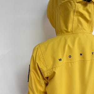 May include: A bright yellow hooded jacket with the word "MONT" printed in black on the back. The jacket has a hood and a black patch on the left sleeve. The jacket is made of a smooth, possibly waterproof material.
