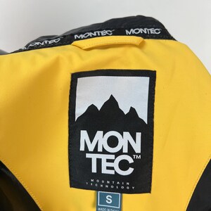 May include: Close-up of a black and yellow jacket collar. The jacket features the brand name "MONTEC" in white on the collar and a large label with the brand logo and "MON TEC MOUNTAIN TECHNOLOGY" text. A size "S" tag is visible.