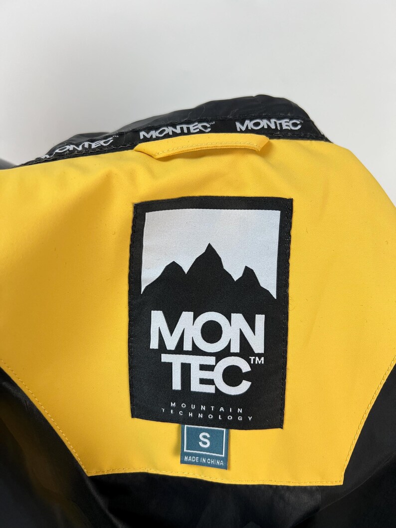 May include: Close-up of a black and yellow jacket collar. The jacket features the brand name "MONTEC" in white on the collar and a large label with the brand logo and "MON TEC MOUNTAIN TECHNOLOGY" text. A size "S" tag is visible.