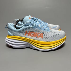 Könnte beinhalten: Paar hellblaue und graue Laufschuhe mit orangefarbenem "HOKA"-Schriftzug. Die Sohle hat eine dicke gelbe Schicht mit weißen und roten Akzenten. Die Schnürsenkel sind hellblau. Diese Schuhe sind für das Laufen und andere sportliche Aktivitäten konzipiert.