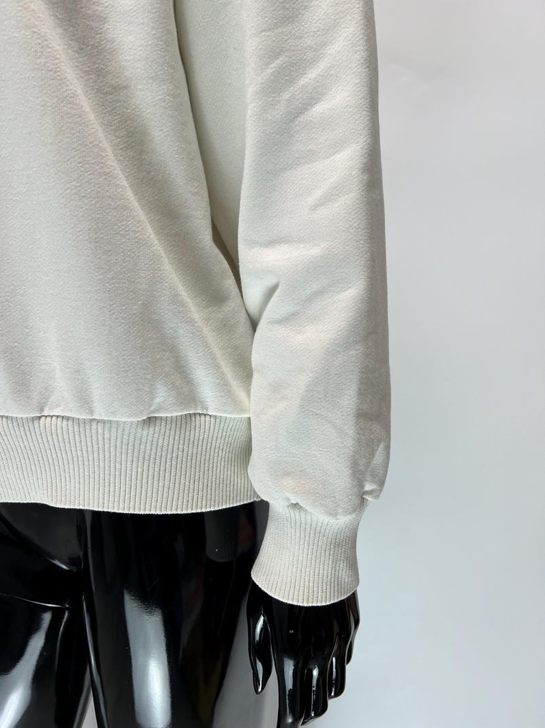 May include: Cream-colored sweatshirt with ribbed cuffs and hem. The long sleeves are gathered at the wrist. The fabric has a textured appearance. The sweatshirt is displayed on a black mannequin.