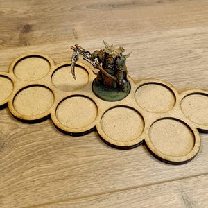 Puede incluir: Un soporte de madera para figuras en miniatura con ocho ranuras circulares. Una figura en miniatura pintada y detallada se encuentra en una ranura. El soporte es de madera marr&oacute;n claro con bordes marrones oscuros.