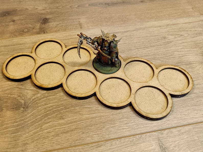 Puede incluir: Un soporte de madera para figuras en miniatura con ocho ranuras circulares. Una figura en miniatura pintada y detallada se encuentra en una ranura. El soporte es de madera marr&oacute;n claro con bordes marrones oscuros.